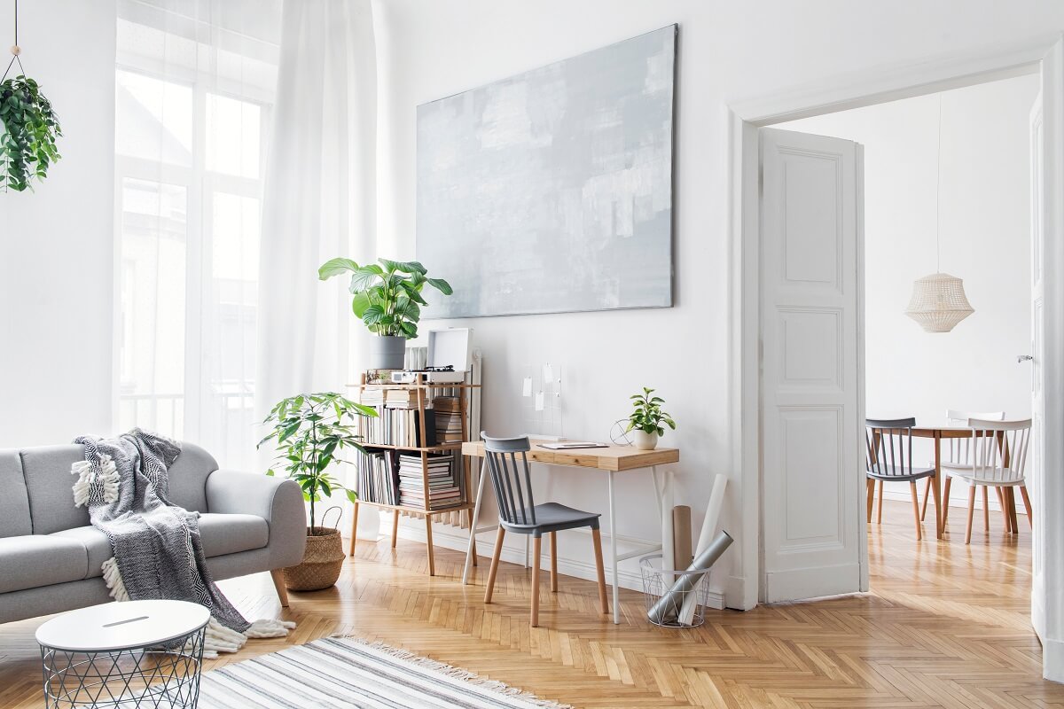 Jasny salon w stylu skandynawskim z szarą sofą, podłogą w jodełkę i drewnianym biurkiem