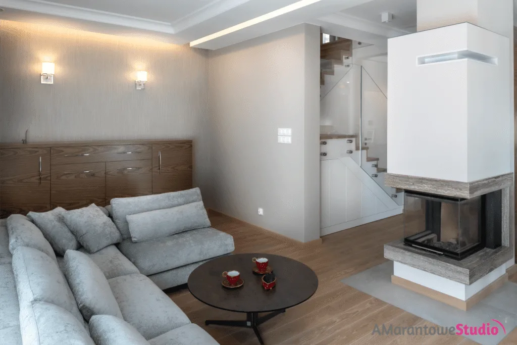 Salon z szarą narożną sofą, okrągłym stolikiem i kominkiem