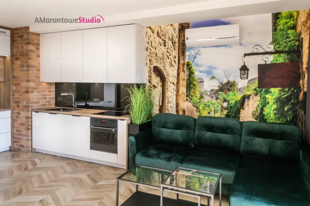 Zielona welurowa sofa przed fototapetą z uliczką i białą kuchnią