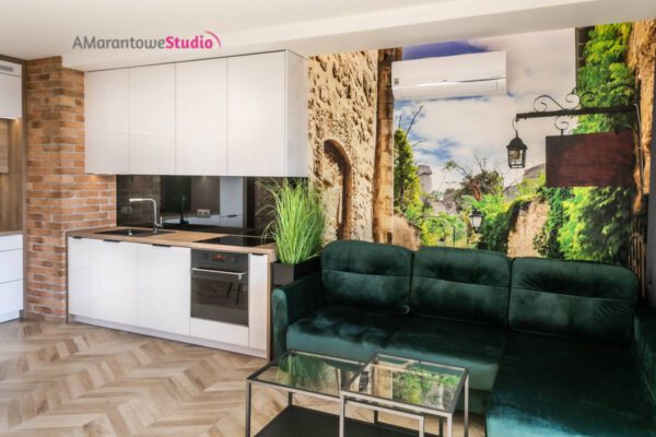 Zielona welurowa sofa przed fototapetą z uliczką i białą kuchnią