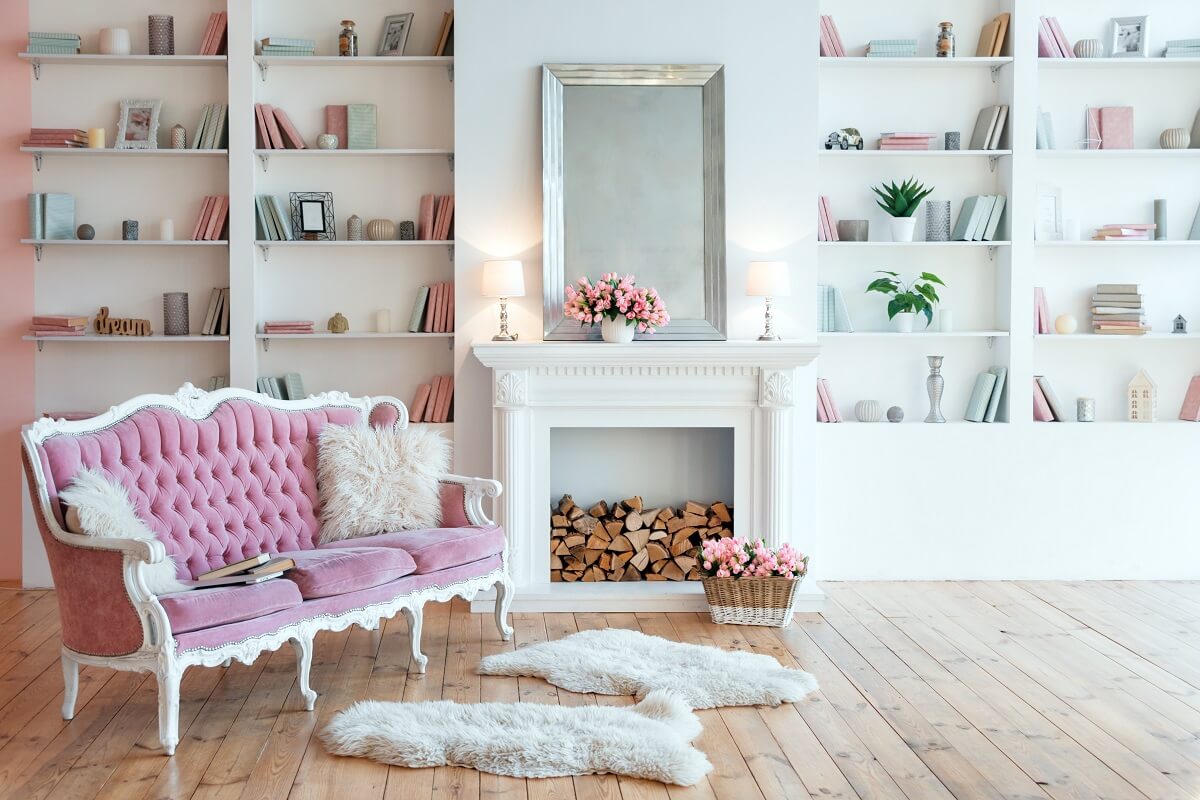 Różowa pikowana sofa, biały kominek i półki z książkami we wnętrzu w stylu francuskim