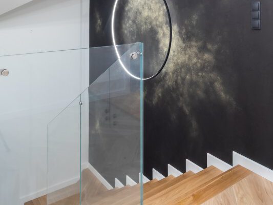 Dębowe schody ze szklaną balustradą przy czarnej ścianie ze złotem