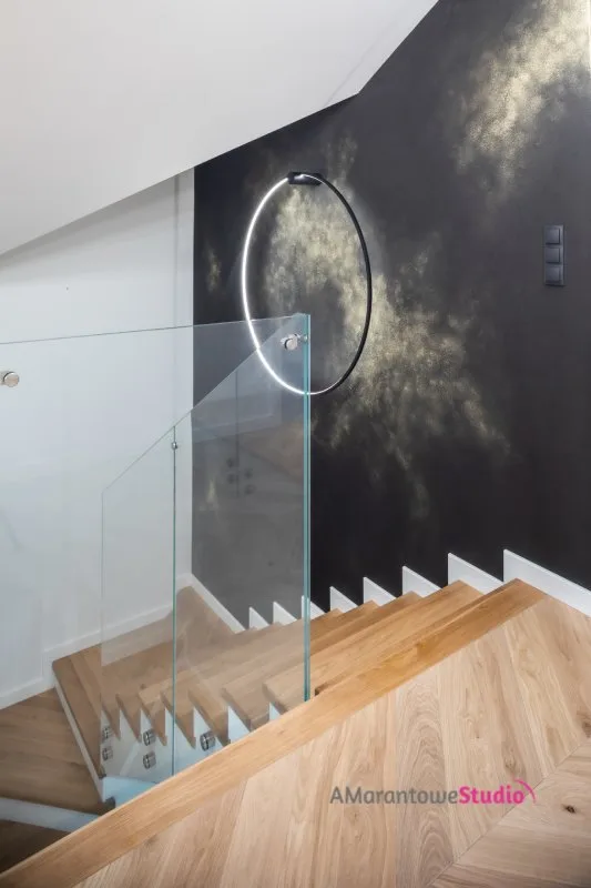 Dębowe schody ze szklaną balustradą przy czarnej ścianie ze złotem